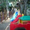 Area giochi recintata, con scivolo colorato e casetta in plastica, tra alberi.