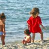 Tre bambini giocano sulla spiaggia vicino all'acqua; uno indossa una maglietta rossa.