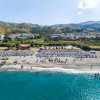 Spiaggia affollata con ombrelloni colorati, mare turchese e colline sullo sfondo.