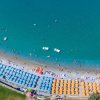 Spiaggia affollata vista dall'alto, con ombrelloni blu e arancioni lungo la costa sabbiosa.