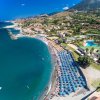 Vista aerea di una spiaggia affollata, ombrelloni allineati e montagne sullo sfondo.